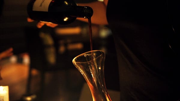 Wine being poured slowly from one glass carafe into a second carafe on a plain kitchen counter