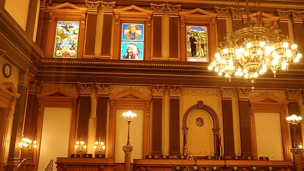 Long wooden hearing benches facing a raised dais in an empty public meeting room lit by ceiling panels