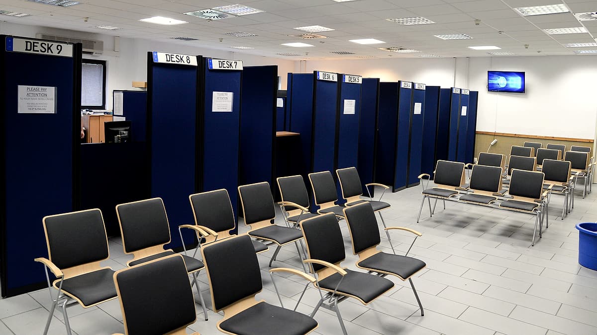 A line of applicants waiting on plastic chairs in a plain corridor, clipboards resting on their knees.